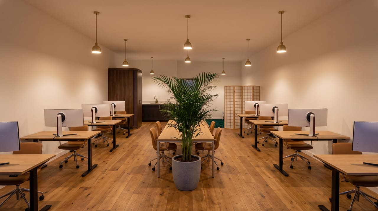 The floor at dusk: two rows of oak standing desks with curved ultrawide monitors under brass pendants, a long shared table with a potted palm down the center, and the musalla's emerald rug behind a folding shoji screen at the far end.