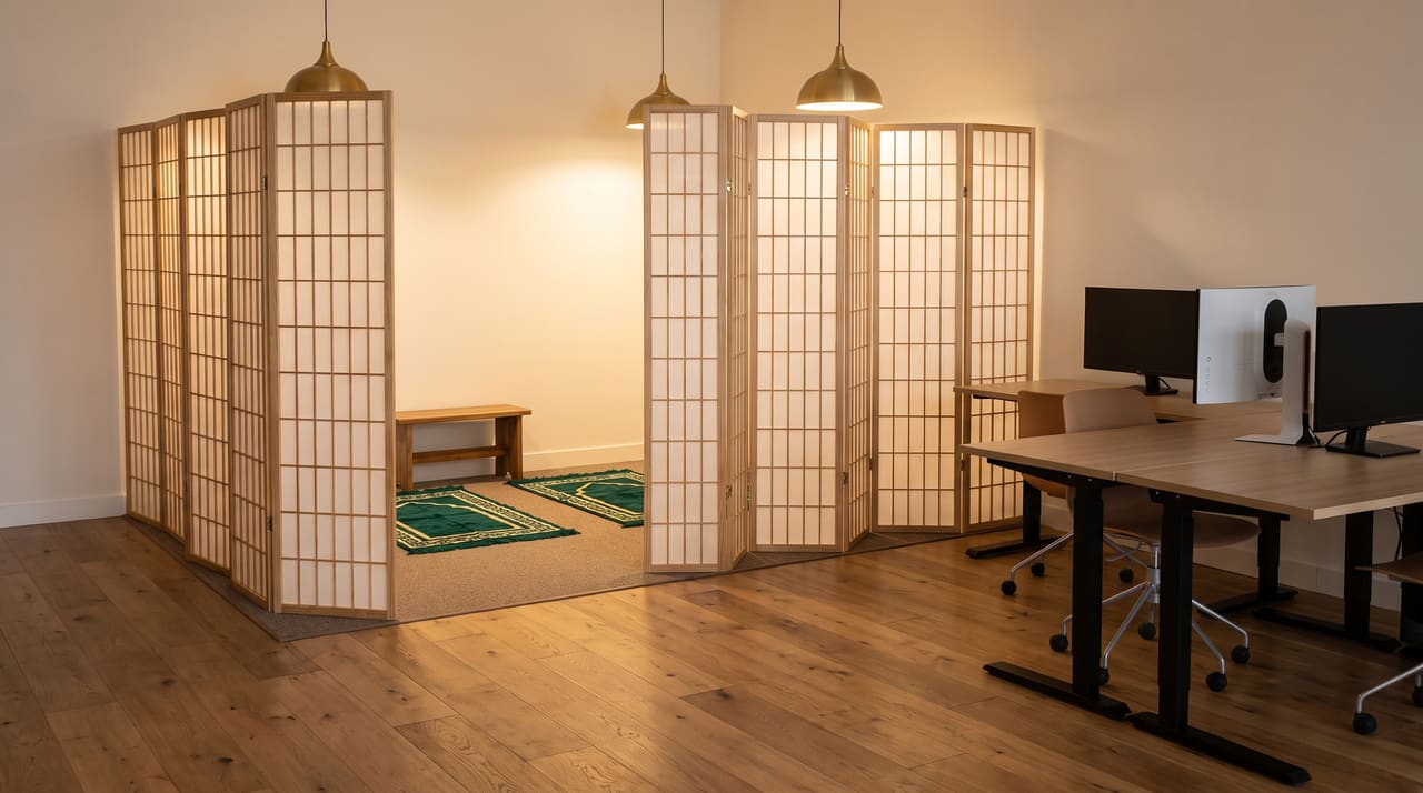 The musalla behind its folding shoji screens: two emerald prayer rugs on carpet, a small wooden bench, warm light over the screens, desks a few steps away.